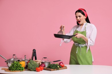 Emotional housewife with pan, spatula and products on pink background. Space for text