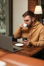 Male blogger with cup of coffee using phone at table in cafe