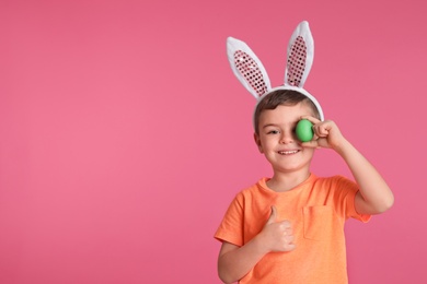 Little boy in bunny ears headband holding Easter egg near eye on color background, space for text
