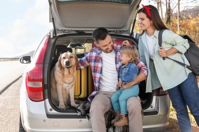 Photo of Parents, their daughter and dog sitting in car trunk outdoors. Family traveling with pet