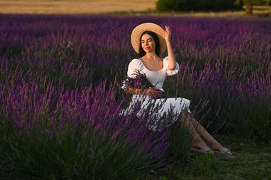 Beautiful young woman with bouquet sitting in lavender field at sunset