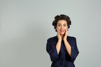 Happy young woman in silk bathrobe with hair curlers on grey background, space for text