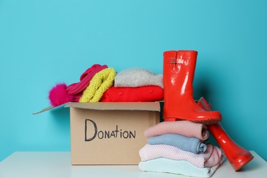 Donation box, knitted clothes and rubber boots on table against color background