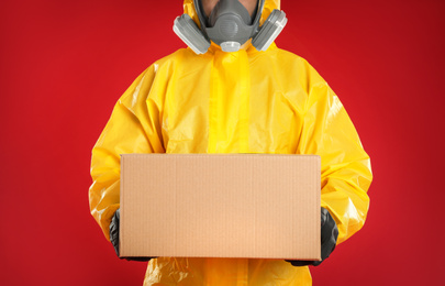 Man wearing chemical protective suit with cardboard box on red background, closeup. Prevention of virus spread