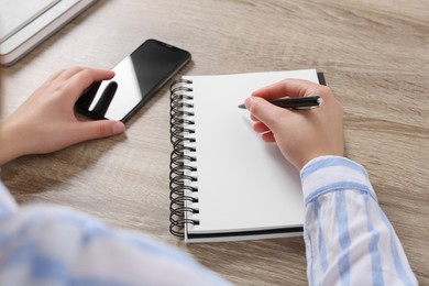 Woman with smartphone writing in notebook at wooden table, closeup