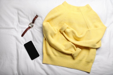 Yellow warm sweater with smartphone and wristwatch on bedsheet, flat lay
