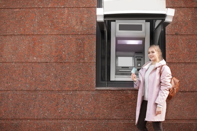 Young woman with credit card near cash machine outdoors. Space for text