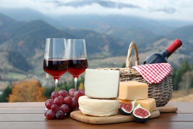 Different types of delicious cheeses, snacks and wine on wooden table against mountain landscape