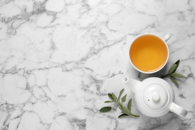 Cup of sage tea, green leaves and teapot on white marble table, flat lay. Space for text