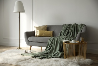 Stylish room interior with comfortable sofa, knitted blanket and coffee table near white wall
