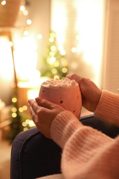 Woman holding cup of hot drink with marshmallows indoors, closeup. Magic Christmas atmosphere