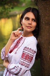 Beautiful woman in embroidered shirt near tree outdoors. Ukrainian national clothes