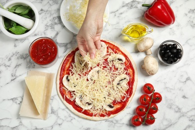 Woman adding grated cheese to unbaked pizza on marble table, top view