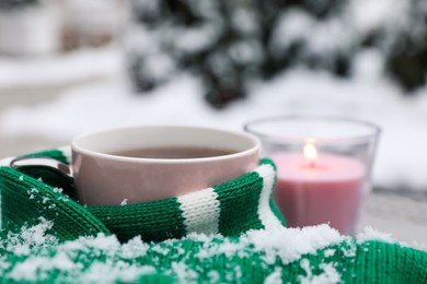 Photo of Winter morning. Cup with hot drink wrapped in green sweater and burning candle outdoors, closeup. Space for text