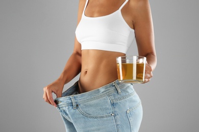 Young woman in old big jeans with cup of tea showing her diet results on grey background, closeup