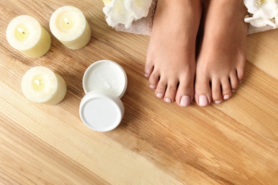Woman with beautiful feet, candles, flowers and moisturizing cream on wooden floor, top view. Spa treatment