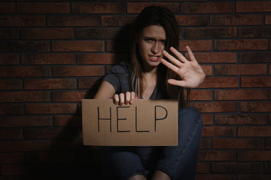 Abused young woman with sign HELP making stop gesture near brick wall. Domestic violence concept