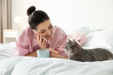 Young woman with cup of coffee talking on phone while lying near cute cat in bedroom. Pet and owner