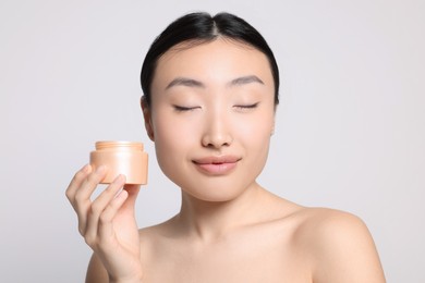 Photo of Beautiful young Asian woman holding jar of body cream on light grey background