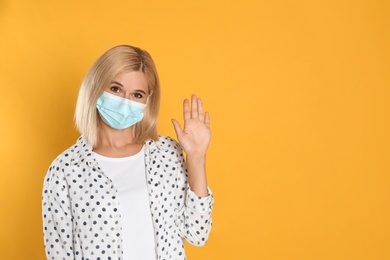 Woman in protective mask showing hello gesture on yellow background, space for text. Keeping social distance during coronavirus pandemic