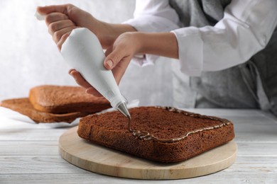 Photo of Woman applying chocolate cream with pastry bag onto homemade sponge cake at white wooden table, closeup