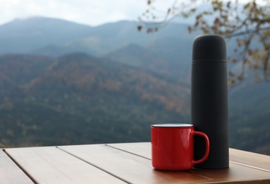 Black thermos and metal mug on wooden table against mountain landscape. Space for text
