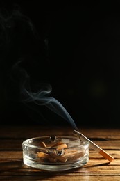 Glass ashtray with stubs and smoldering cigarette on wooden table against black background. Space for text