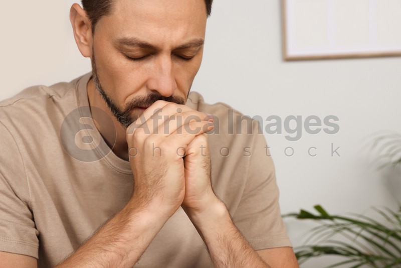 Man with clasped hands praying in room at home, closeup