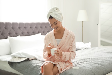 Pretty young woman with towel on head using skin cream in bedroom