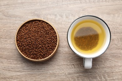 Buckwheat tea in cup and bowl with granules on wooden table, flat lay