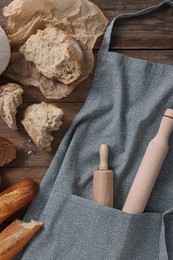 Clean kitchen apron with rolling pins and different types of bread on wooden table, flat lay