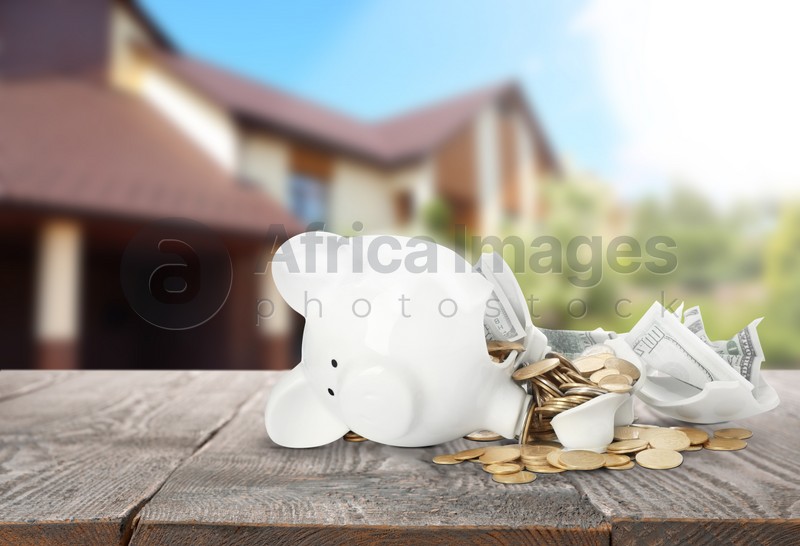 Broken piggy bank with money on wooden surface and blurred view of beautiful house. Mortgage concept