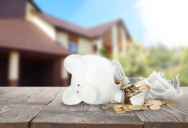 Broken piggy bank with money on wooden surface and blurred view of beautiful house. Mortgage concept