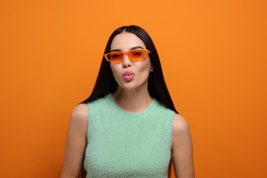 Photo of Beautiful young woman in stylish sunglasses blowing kiss on orange background