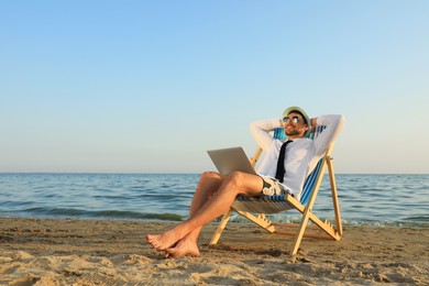 Happy man with laptop resting on deckchair near sea. Business trip