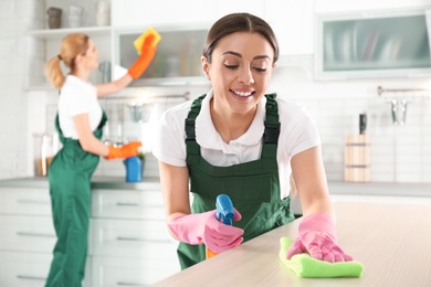 Woman using rag and sprayer for cleaning table with colleague in kitchen