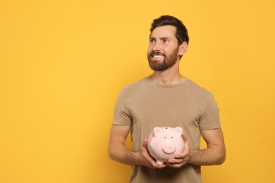 Happy man with ceramic piggy bank on orange background, space for text