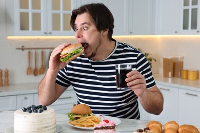Hungry overweight man with tasty burger and glass of cold drink at table in kitchen