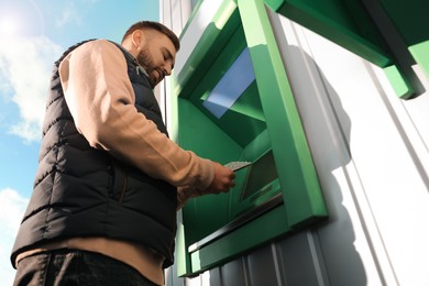 Photo of Young man using cash machine for money withdrawal outdoors, low angle view
