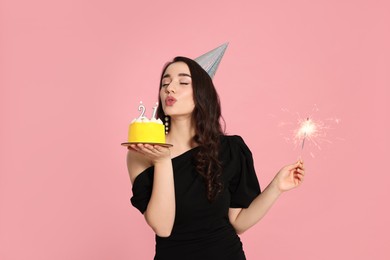 Photo of Coming of age party - 21st birthday. Woman holding delicious cake and sparkler and blowing number shaped candles on pink background