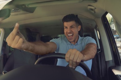 Stressed angry man showing middle finger in modern car, view through windshield
