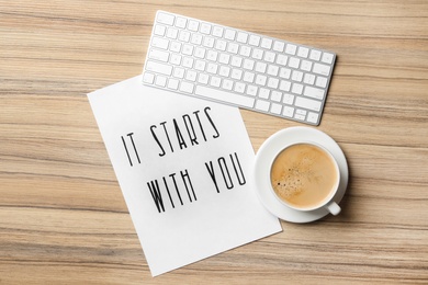 Sheet of paper with phrase It Starts With You, coffee and keyboard on wooden table, flat lay
