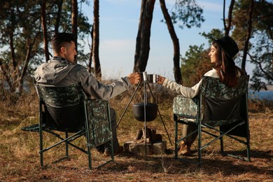 Photo of Couple resting in camping chairs and enjoying hot drink near bonfire outdoors