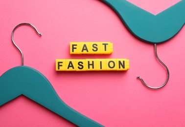 Yellow plastic cubes with phrase Fast Fashion and hangers on pink background, flat lay