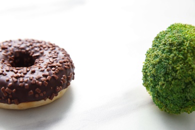Concept of choice between healthy and junk food, closeup. Tasty doughnut with broccoli on white table