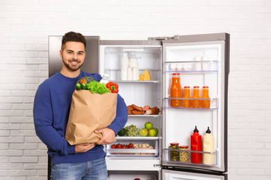 Young man with paper bag full of products near open refrigerator indoors