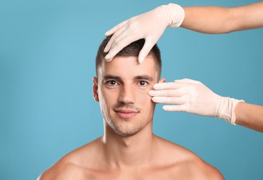 Doctor examining man's face for cosmetic surgery on light blue background