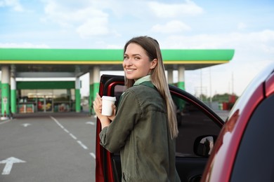 Beautiful young woman with coffee opening car door at gas station