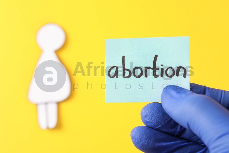 Doctor holding card with word ABORTION and paper cutout of pregnant woman on yellow background, closeup. Space for text