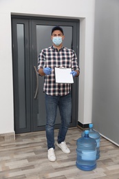 Courier in face mask with clipboard and bottles of cooler water in entryway. Delivery during coronavirus quarantine